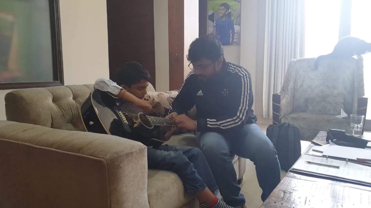 A young Mir Kahol learning acoustic guitar with his teacher on a couch